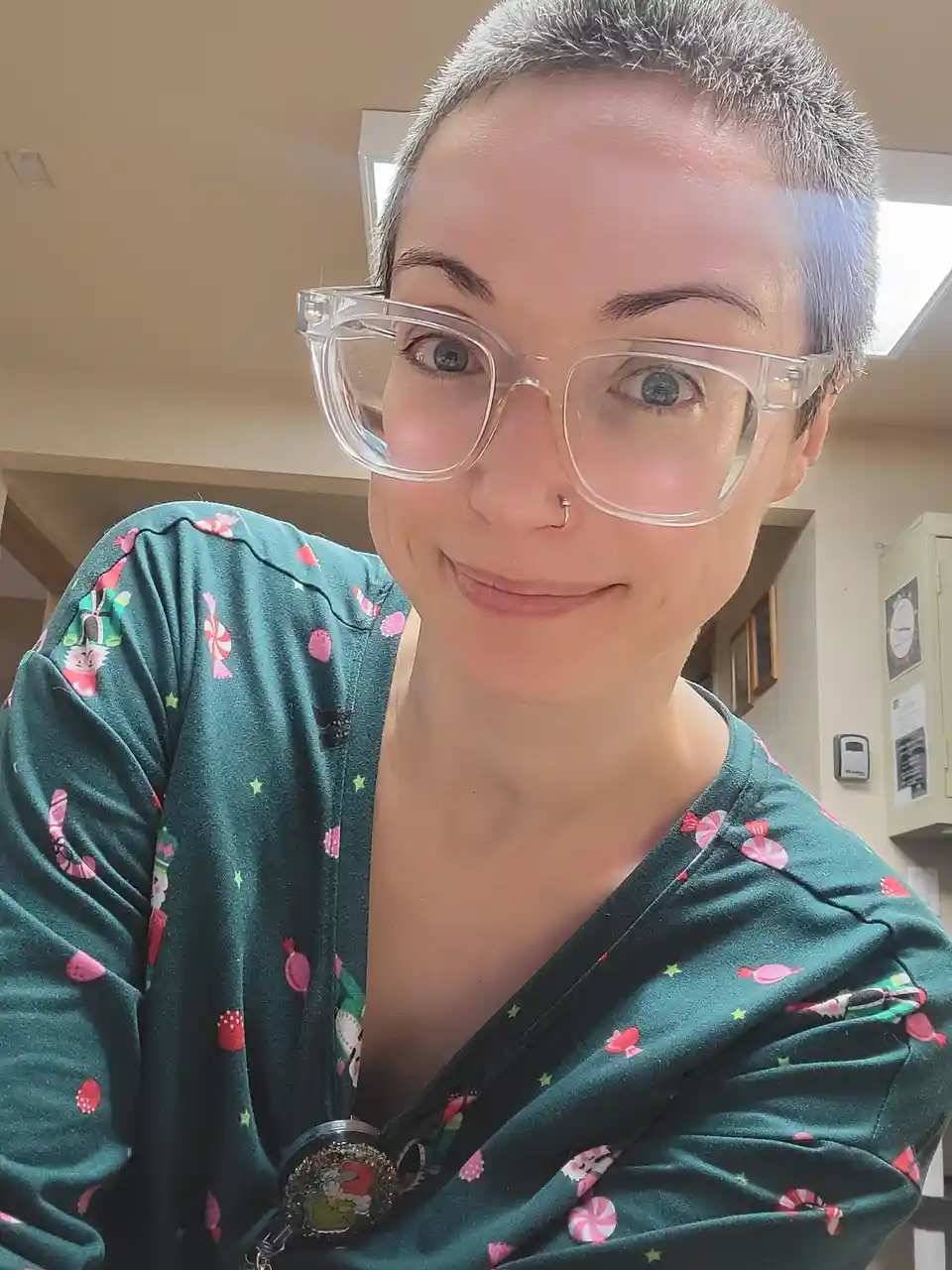 A person with short gray hair and clear-framed glasses smiles at the camera while wearing a dark green scrub top decorated…