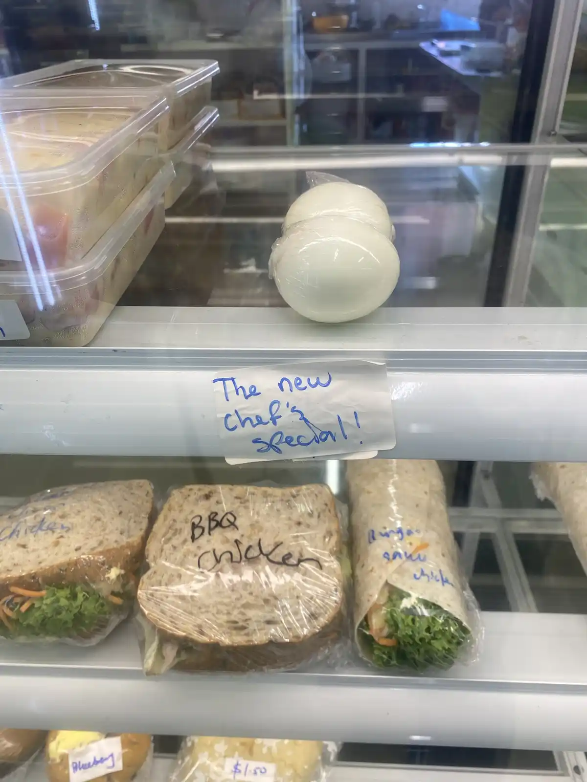 A refrigerated display case shows sandwiches and wraps labeled with handwritten names like 'BBQ Chicken' and 'Burgo sau…