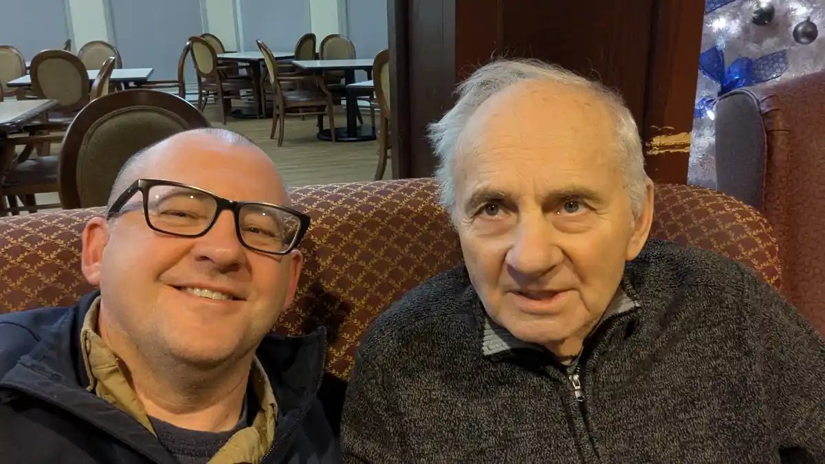 Two men sit side by side on a patterned couch in a restaurant or lounge. The man on the left, wearing glasses and a jacket…