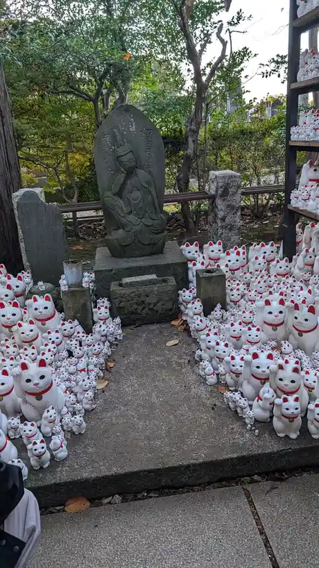 A stone statue of a seated figure is surrounded by hundreds of white ceramic cat figurines, each with red collars and raised…