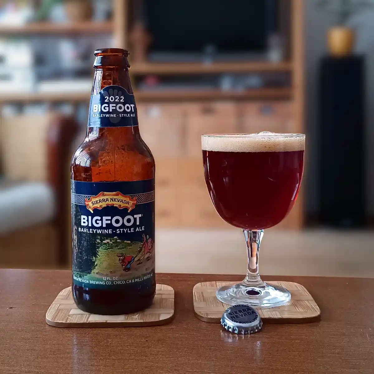 A bottle of 2022 Sierra Nevada Bigfoot Barleywine-Style Ale sits next to a glass filled with the dark red beer, both resting…