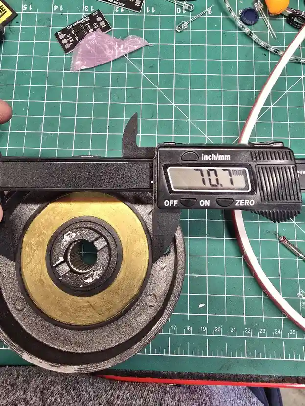 A digital caliper displays the measurement '70.7' while measuring a brass and steel mechanical part on a green cutting mat…