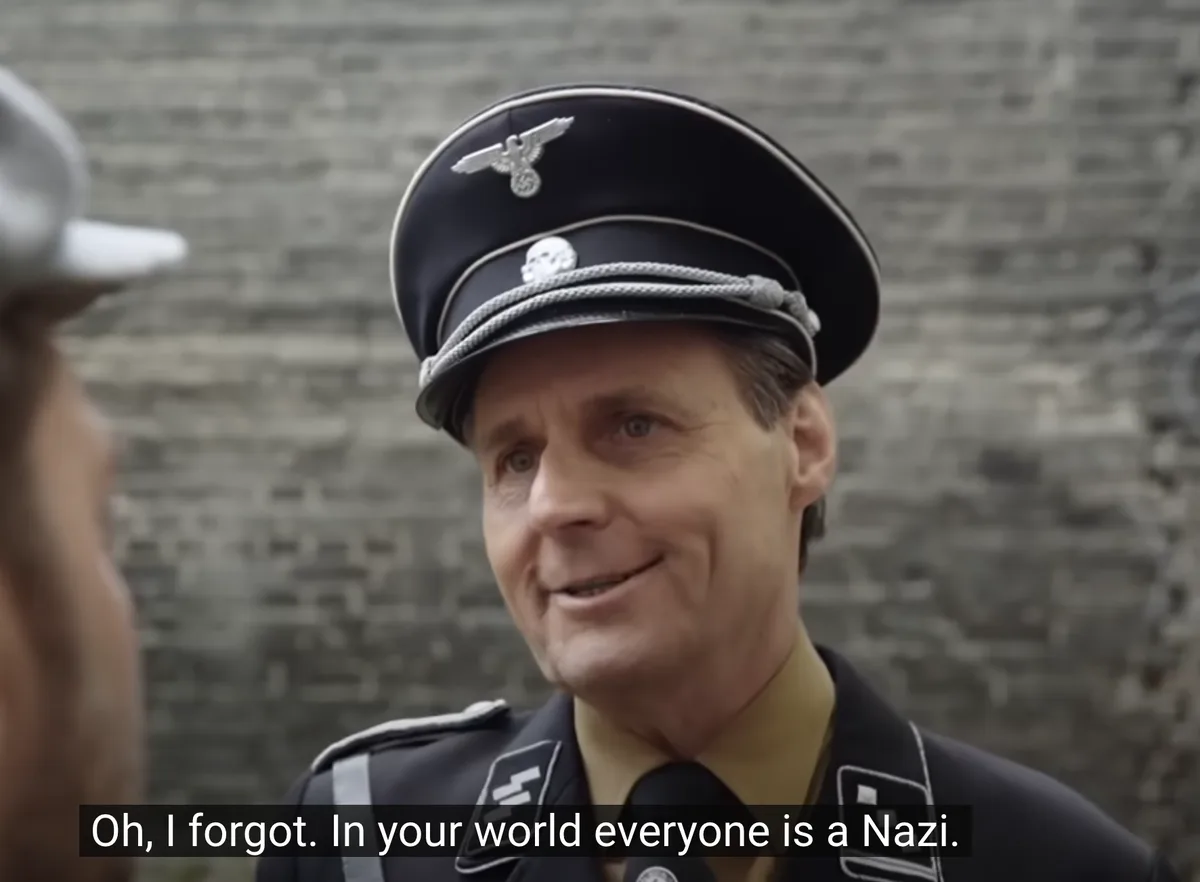 A man in a Nazi uniform with an eagle and swastika emblem on his cap and SS insignia on his collar smiles while speaking to…