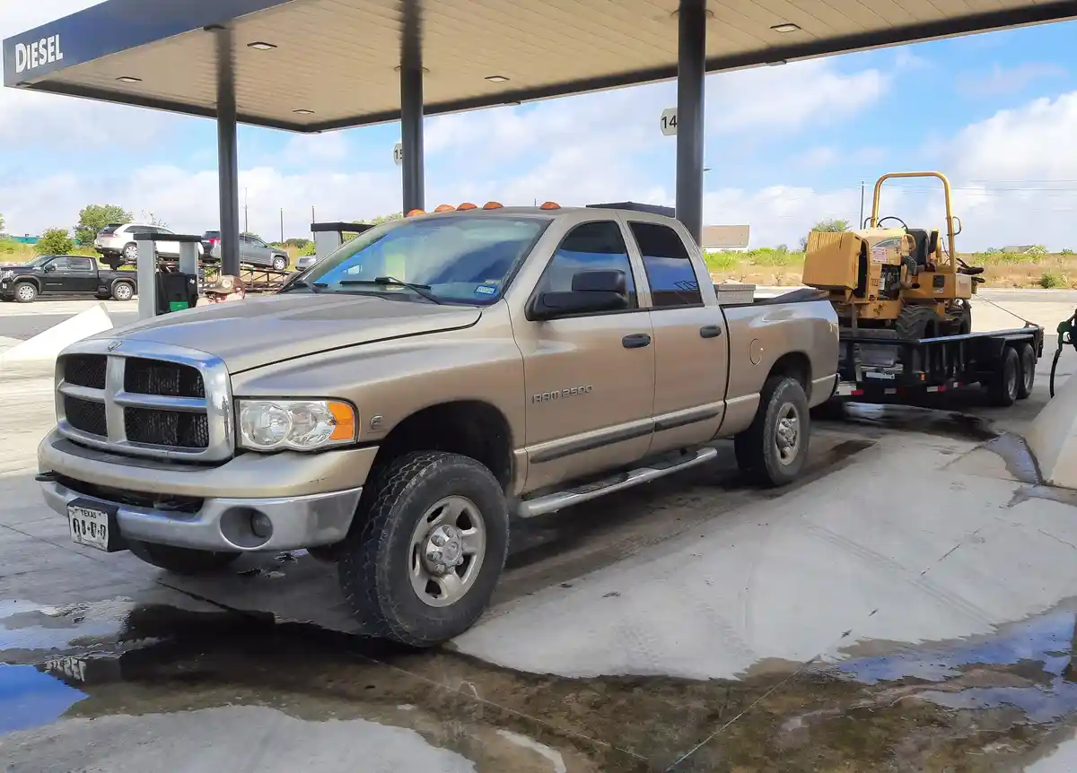 A tan Dodge Ram 2500 pickup truck is parked at a diesel fuel pump, pulling a trailer with a yellow piece of construction…