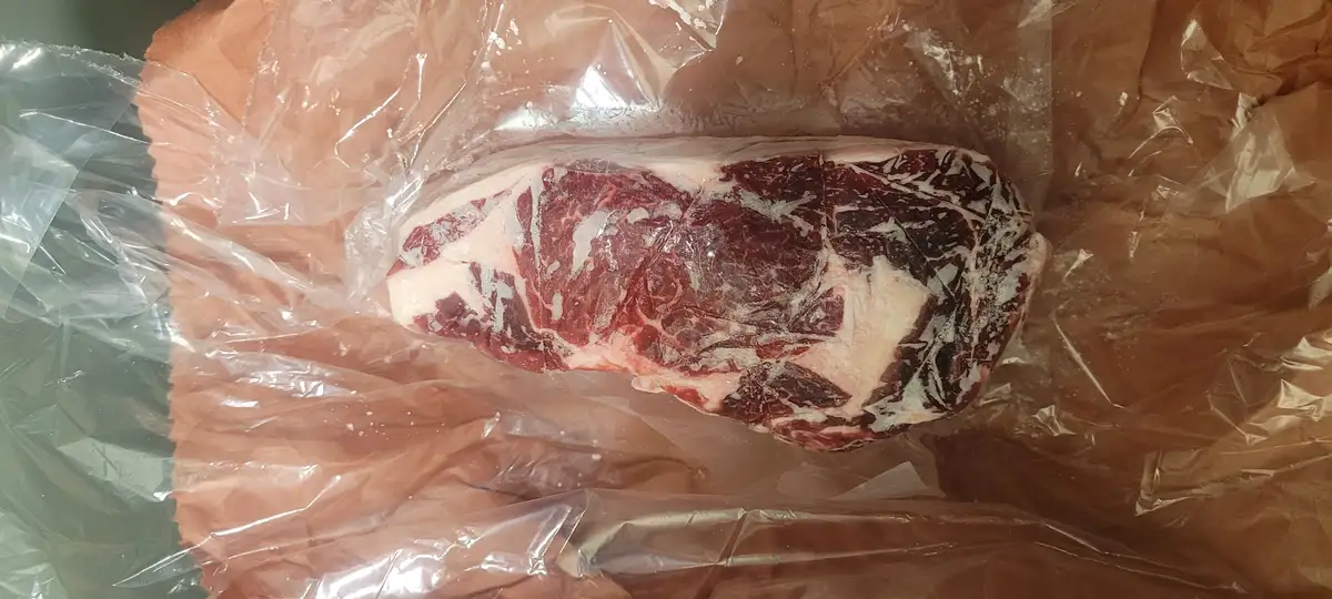A large, raw, marbled beef steak with visible fat and muscle fibers rests inside a crinkled, transparent plastic bag. The…