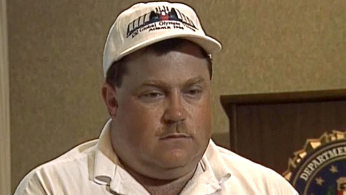 A man wearing a white baseball cap with '1996 Global Olympics Atlanta' text and a white polo shirt looks directly at the…