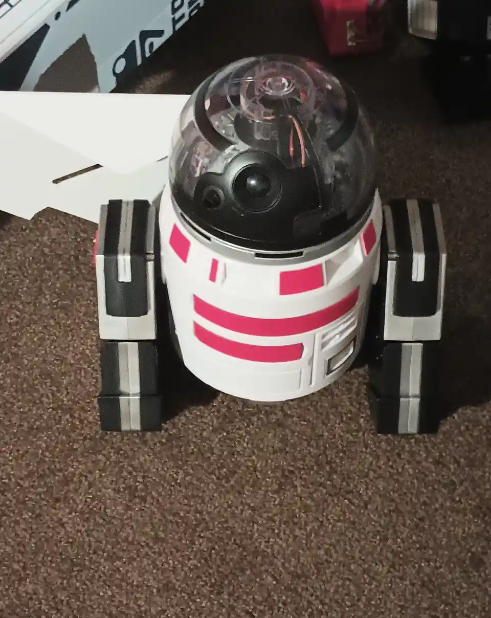 A white and pink droid with black and silver legs sits on a brown carpet, its transparent dome revealing internal wiring and…