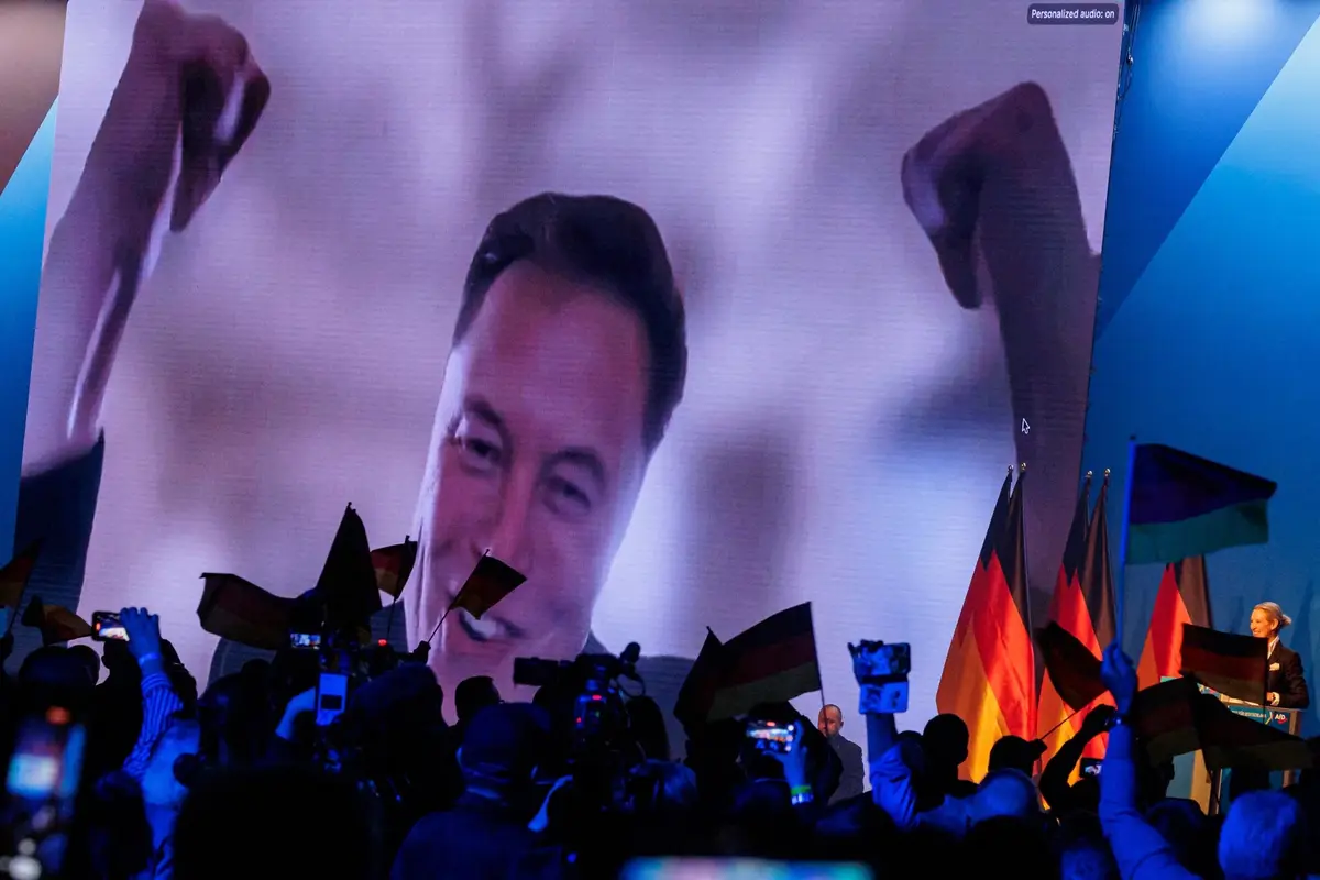 A large screen displays a smiling man with raised fists, viewed from below by a crowd holding German flags and recording on…