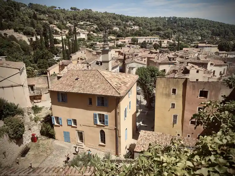 An aerial view of a sun-drenched European village with terracotta roofs, ochre buildings, and lush green hills in the…