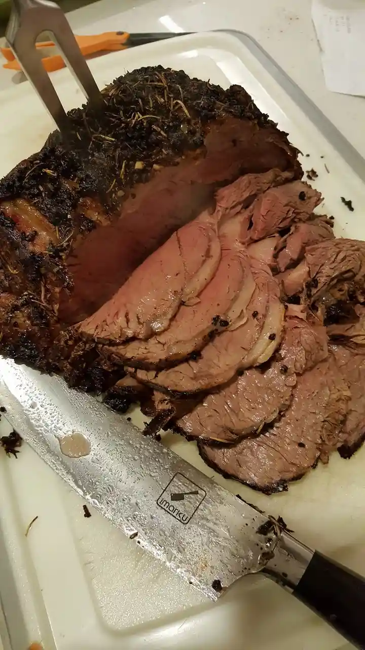 A close-up photo shows a large, roasted beef roast on a white cutting board, partially sliced to reveal pink, juicy interior…