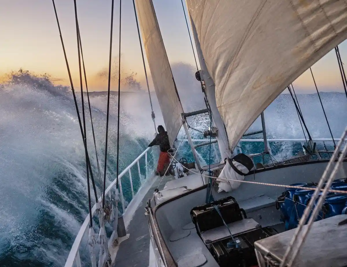 A person in dark clothing and red pants stands on the deck of a sailboat as it cuts through large, foamy ocean waves during…