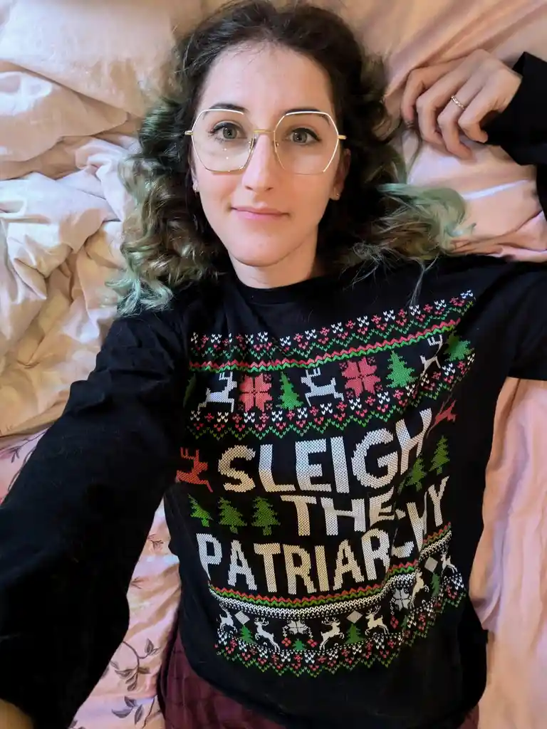 A person with curly hair and glasses lies on a bed wearing a black Christmas sweater with reindeer, trees, and snowflakes…