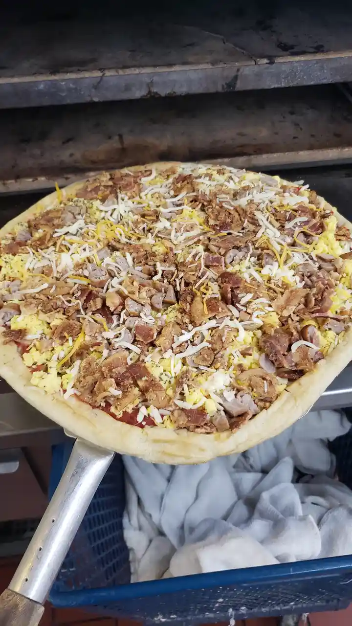A pizza is being held on a metal peel in front of an oven, topped with scrambled eggs, bacon, sausage, and shredded cheese…