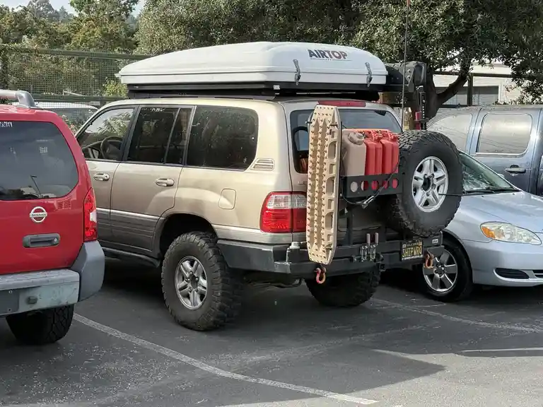 A beige SUV is heavily modified with a rooftop cargo box, a spare tire mounted on the rear, red fuel cans, a ladder, and…