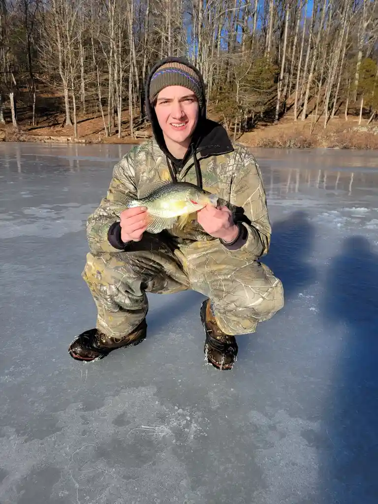 A person in full camouflage winter gear, including a beanie and snow boots, is crouching on a frozen lake holding up a small…