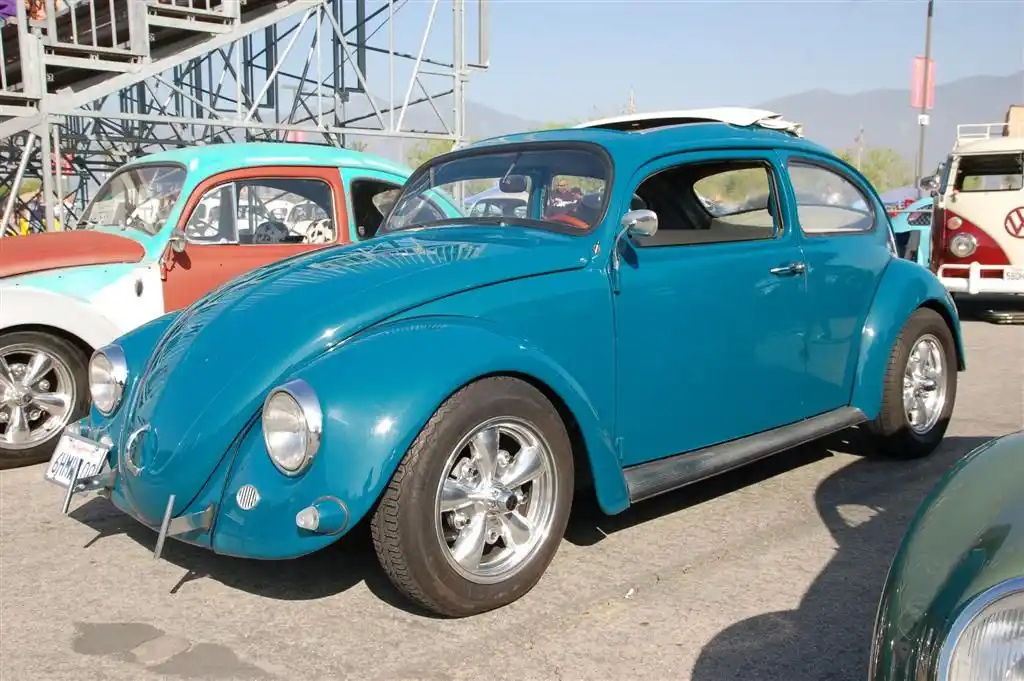 A shiny turquoise Volkswagen Beetle with chrome wheels and a surfboard on its roof is parked at a car show, with other…