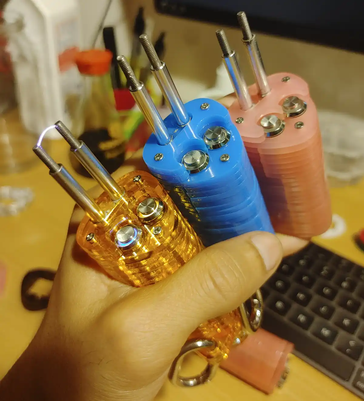 A hand holds three colorful, 3D-printed keychain devices with metal rods protruding from their tops; the orange one in the…