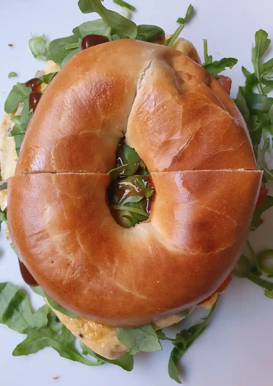 A top-down photo of a bagel sandwich with arugula, sauce, and other fillings, where the bagel has been sliced into four…
