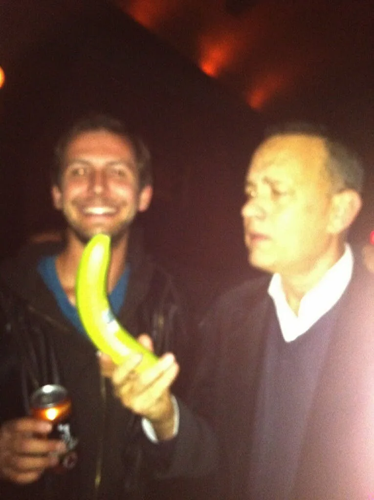 Two men are in a dimly lit, reddish environment, possibly a bar or club. The man on the left, smiling broadly, holds a green…