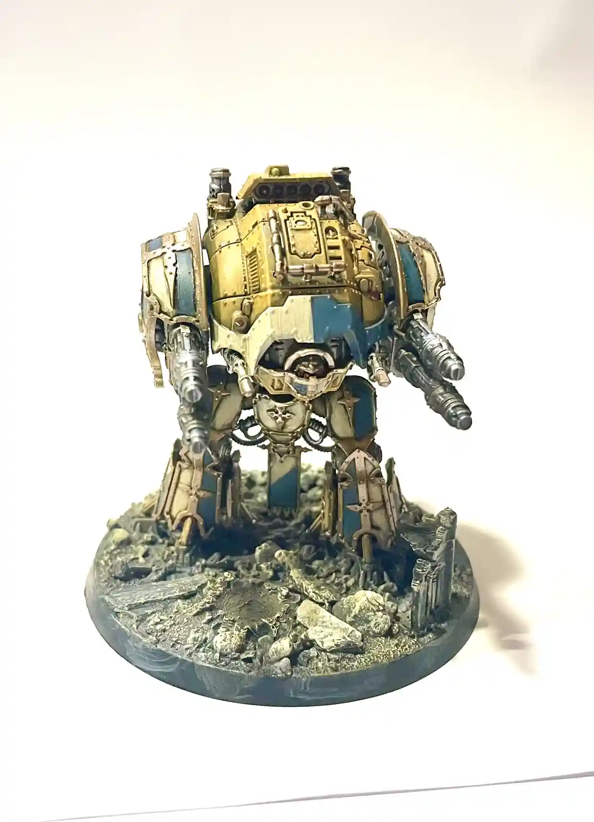 A heavily armored, gold-and-blue painted war machine miniature stands on a rocky, debris-strewn base against a plain white…