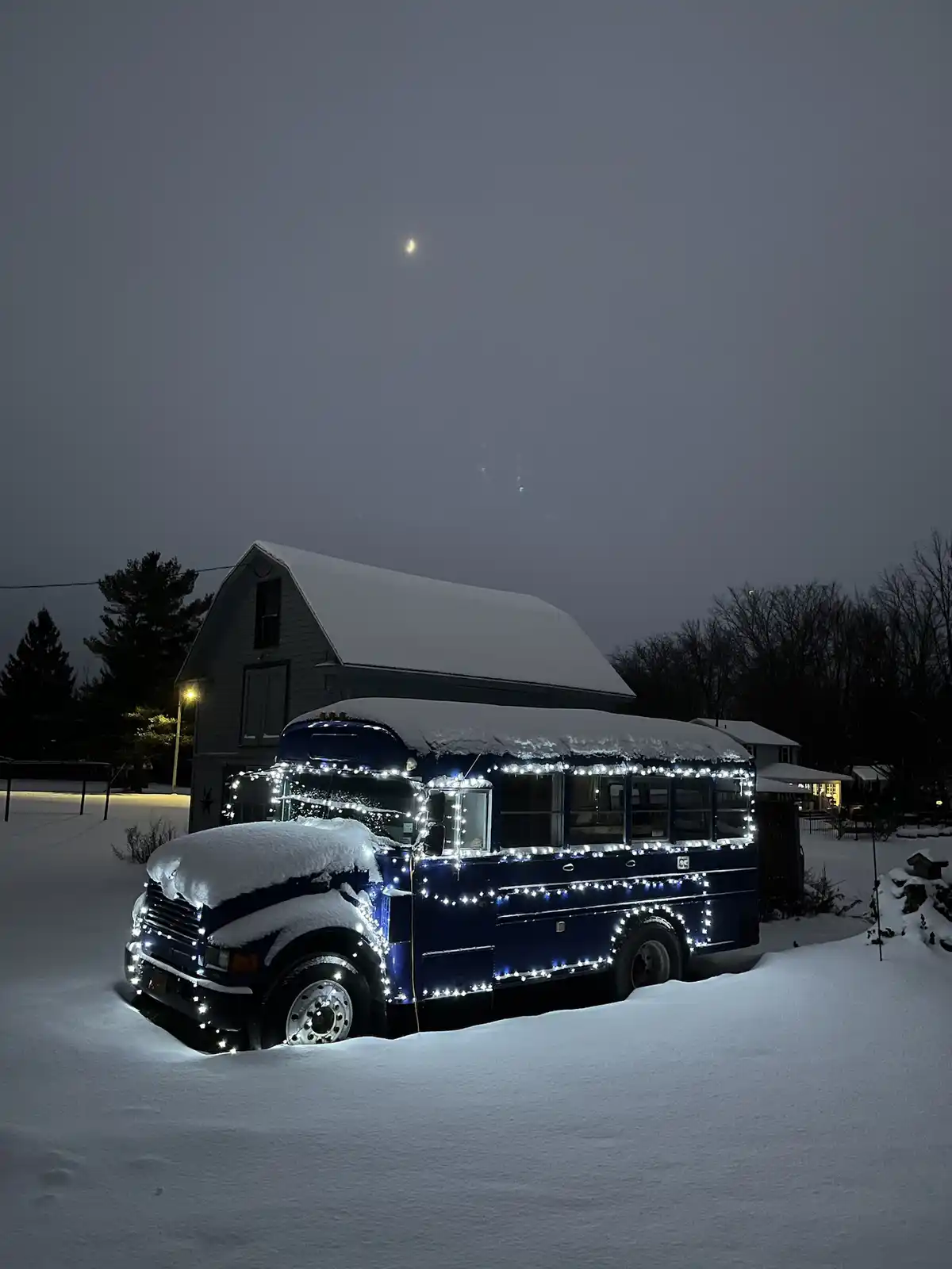 A blue school bus is parked in deep snow at night, covered in snow on its roof and hood, but decorated with bright white…