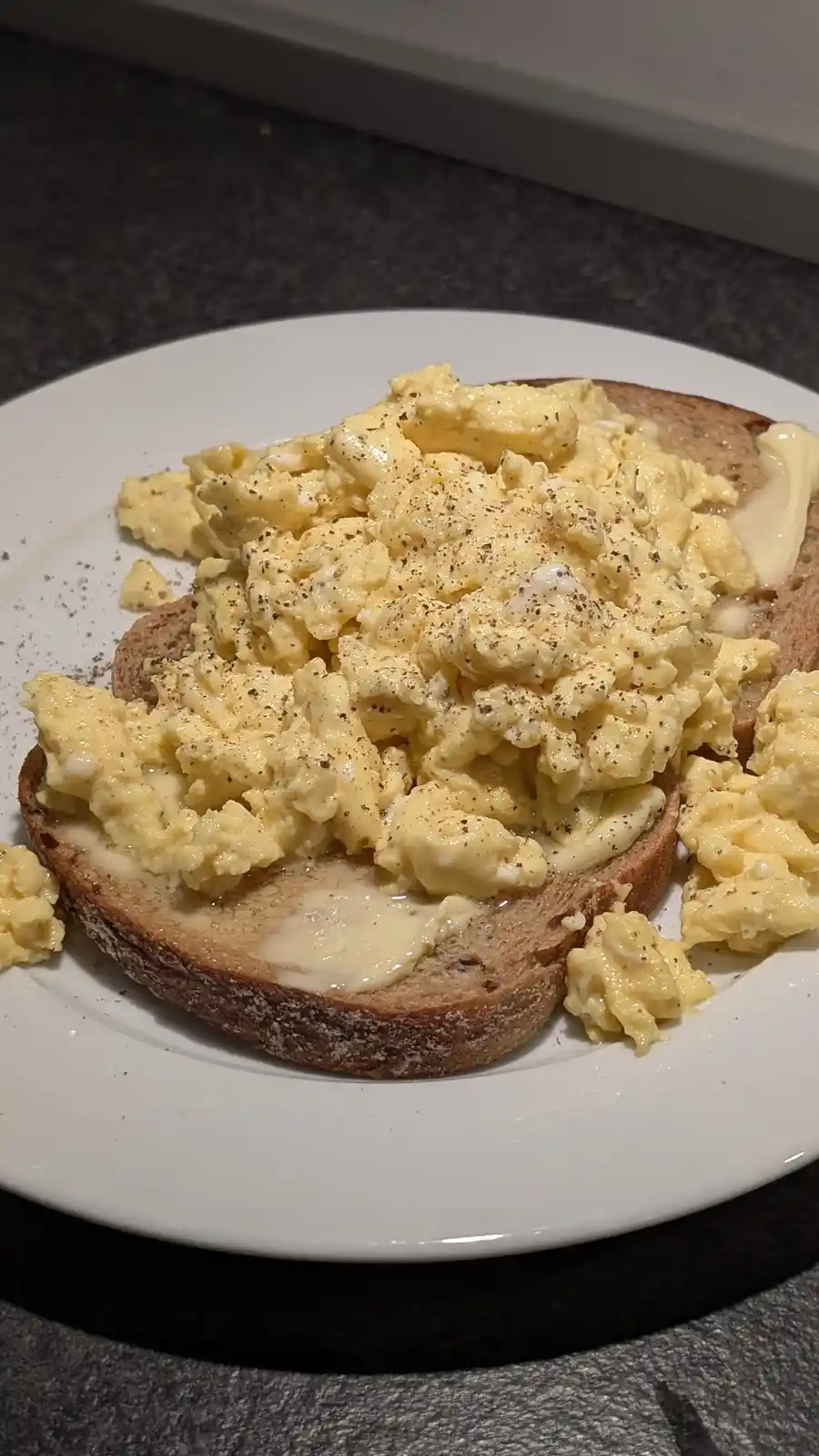 A close-up photo shows a white plate holding two slices of toasted bread, each generously topped with creamy scrambled eggs…