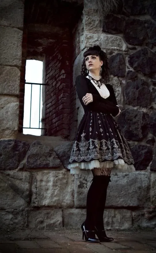 A woman in a black and white gothic lolita dress stands with arms crossed in front of a stone wall with a barred window. She…