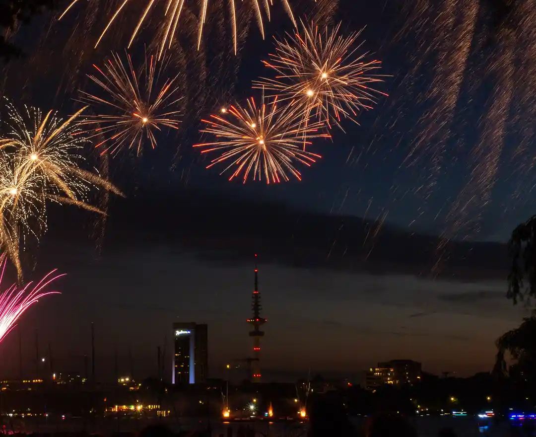 A long-exposure photograph captures vibrant fireworks exploding in the night sky over a cityscape, with a tall tower and…