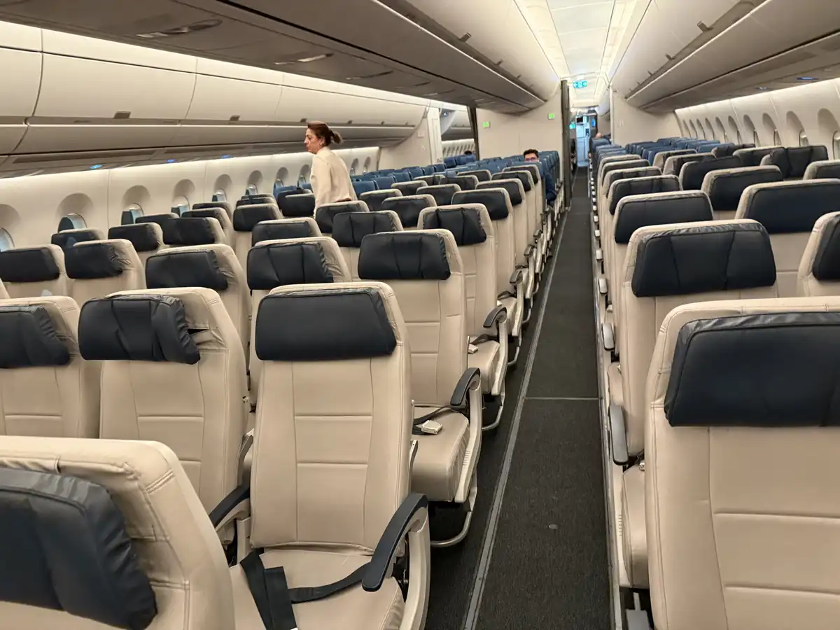 An airplane cabin with rows of empty beige and black seats, a flight attendant walking down the aisle, and one passenger…