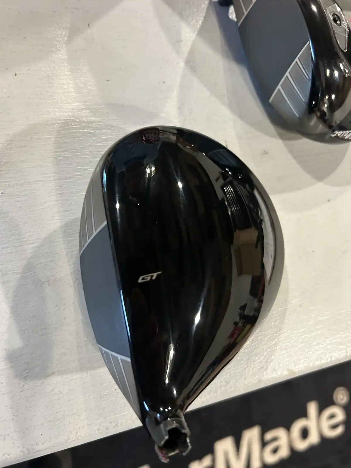 A close-up photo shows a shiny black and gray golf driver head with 'GT' printed on it, resting on a white textured surface…