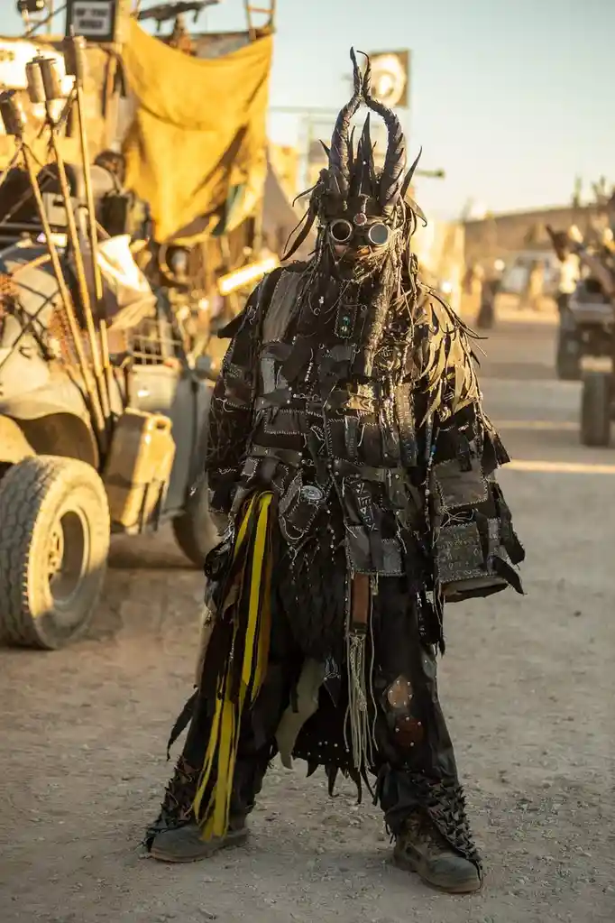 A person in an elaborate, dark, and heavily accessorized costume with a horned helmet and goggles stands in a desert setting…