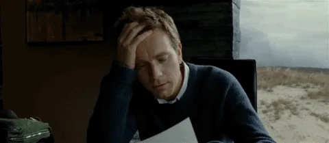 A man in a dark sweater sits at a desk, initially looking downcast and rubbing his forehead while holding a piece of paper…