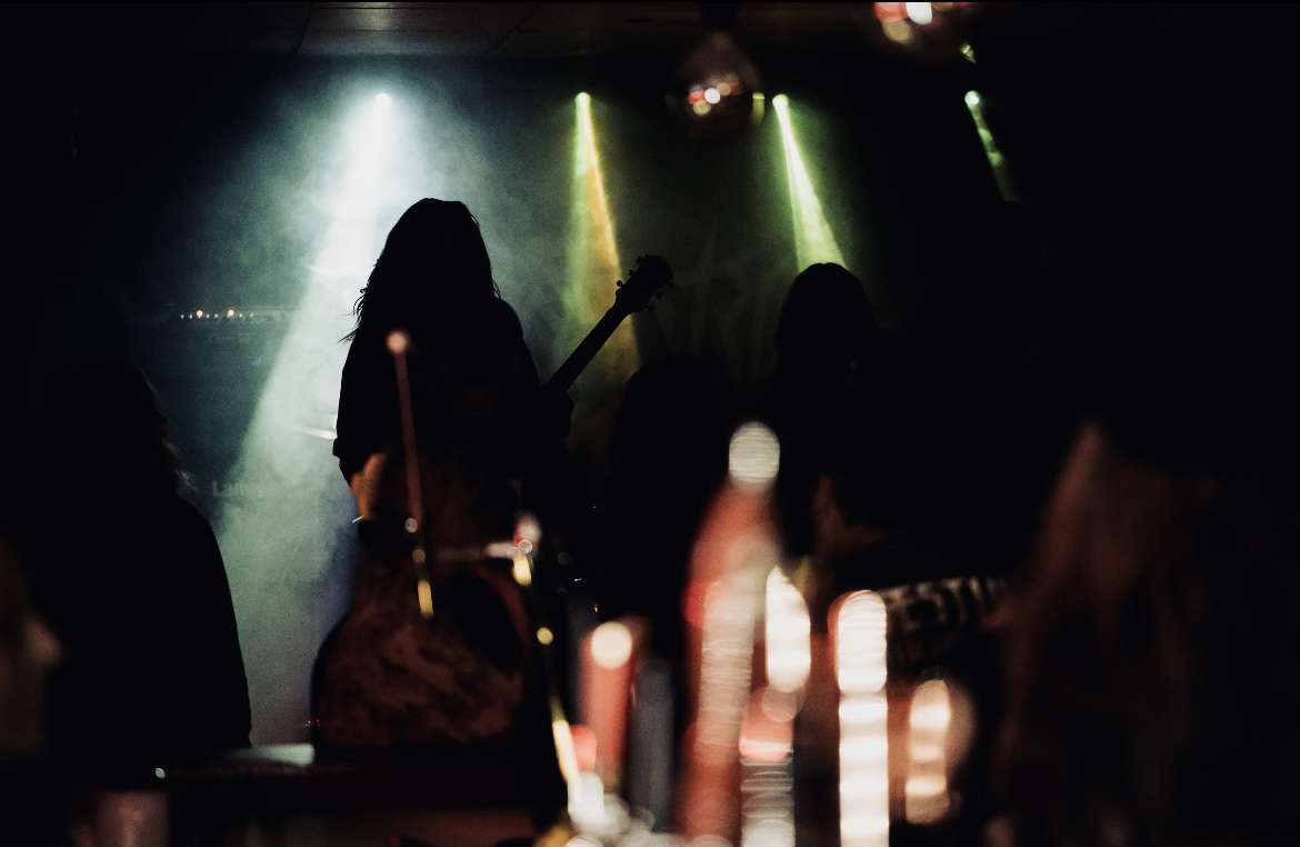 A dimly lit concert photo shows silhouettes of musicians on stage under spotlights, with blurred audience members and…