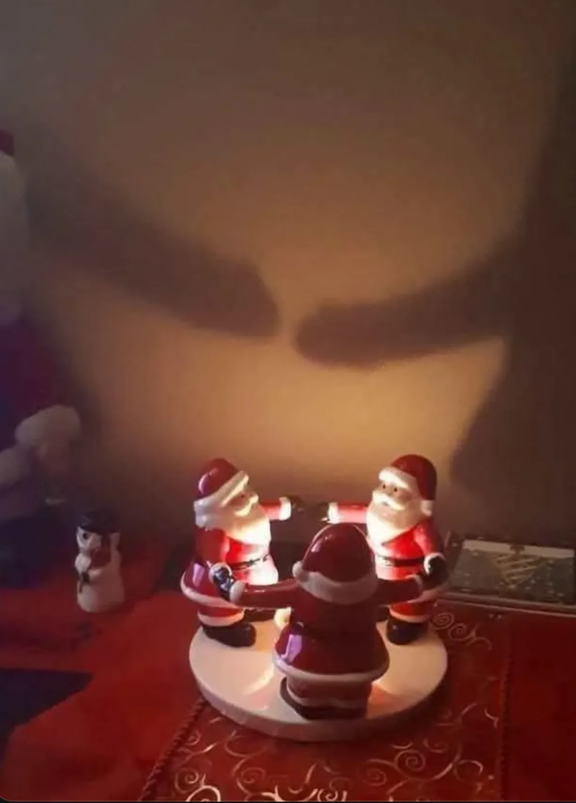 Three illuminated Santa Claus figurines are arranged in a circle on a white base, appearing to hold hands. Their shadows…