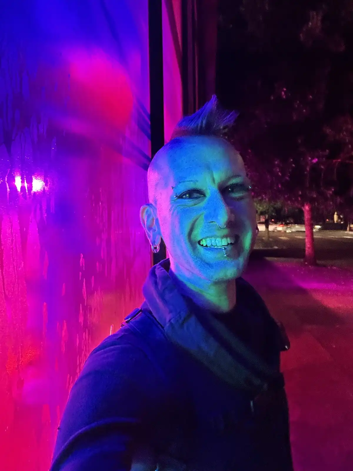 A person with a shaved head and a mohawk stands outside at night, illuminated by vibrant purple and blue neon lights…