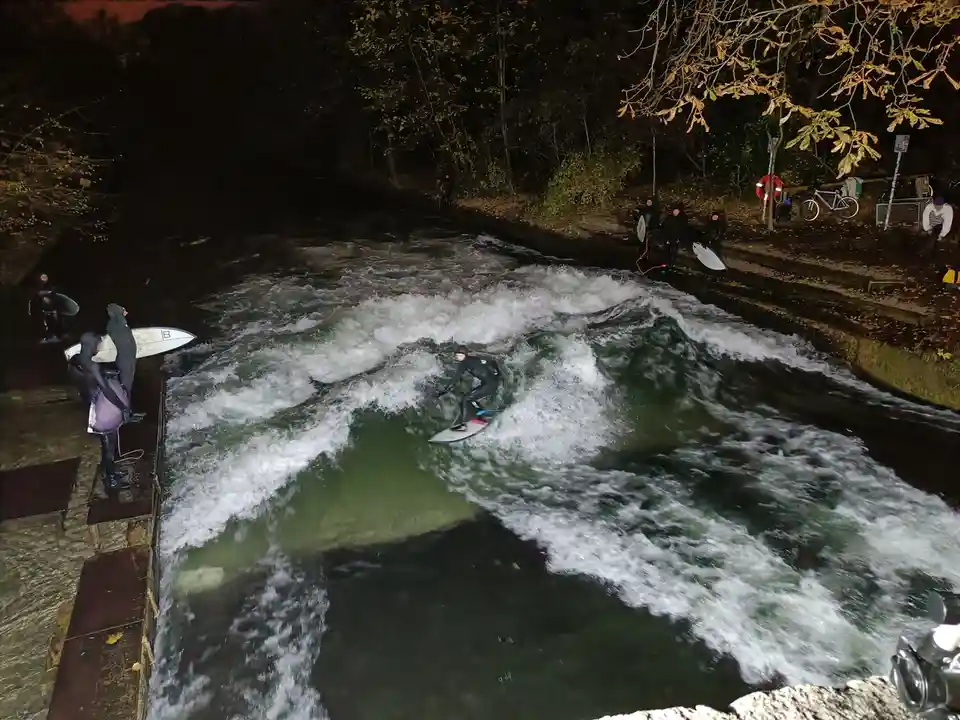 A person in a wetsuit is surfing on a turbulent river at night, surrounded by other people in wetsuits holding surfboards on…