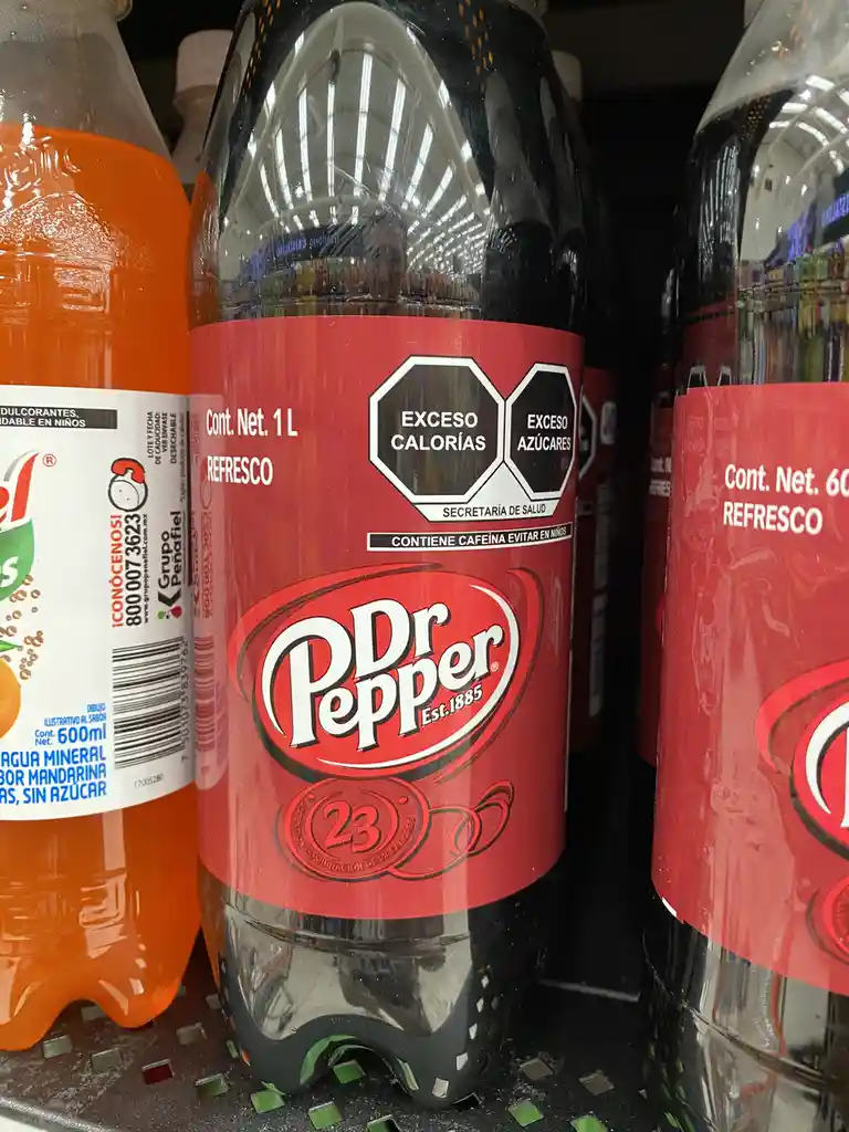 A close-up photo of a 1L Dr Pepper 23 soda bottle on a store shelf, showing its red label with health warnings in Spanish…