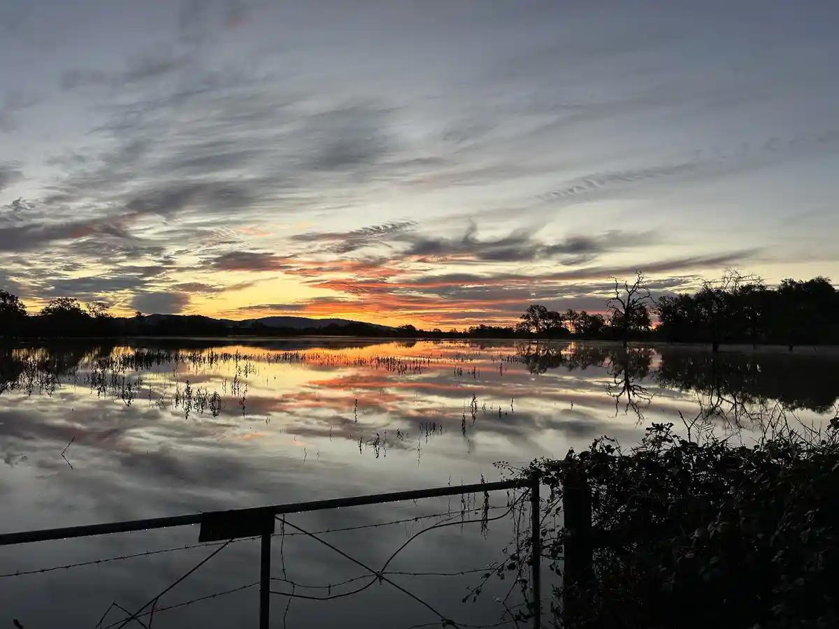 A serene sunset over a flooded field with trees and hills in silhouette, their reflections mirrored in the water, viewed…