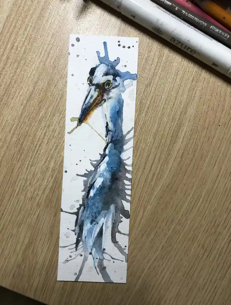 A watercolor painting on a white bookmark depicts a bird with a long beak and blue-gray feathers, but its head and neck are…