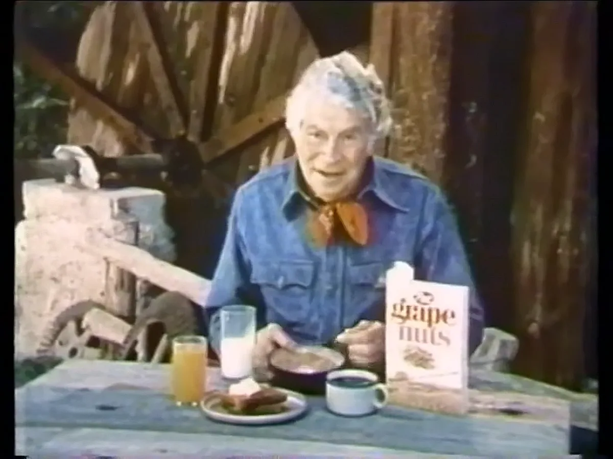 An elderly man with white hair, wearing a blue denim shirt and orange bandana, sits at a wooden table outdoors. He is eating…