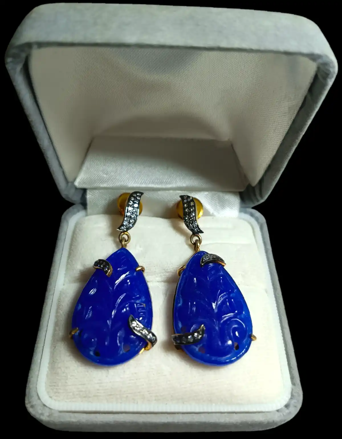 A pair of ornate, deep blue carved stone earrings with gold and diamond accents sit in an open white velvet jewelry box. The…