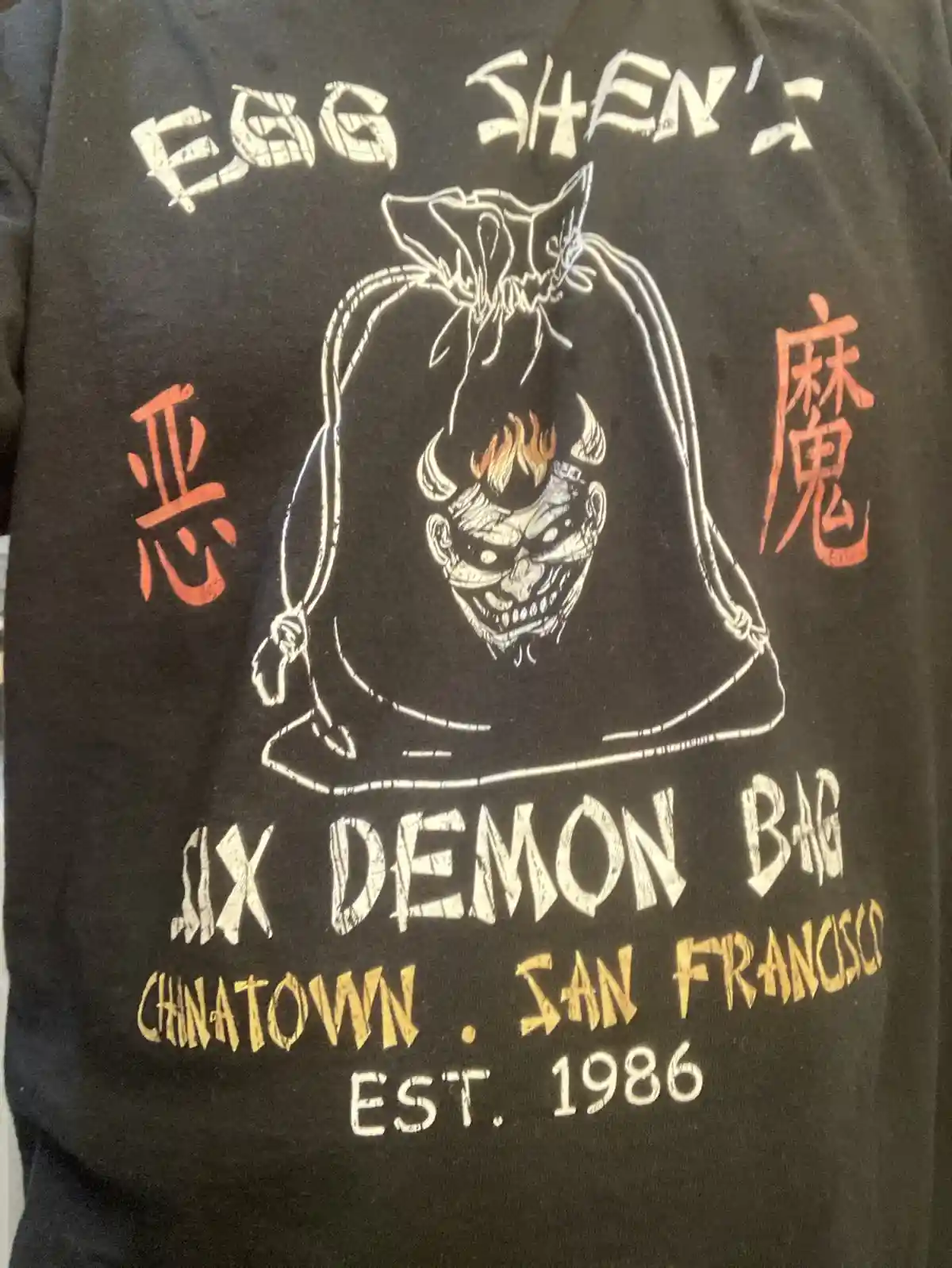 A black t-shirt features a white outline of a drawstring bag with a demonic face inside, flanked by red Chinese characters…
