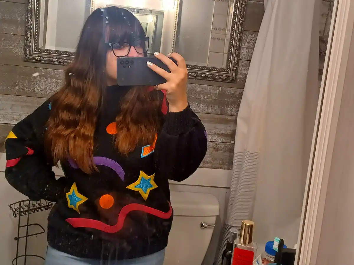 A person with long brown hair and glasses takes a mirror selfie in a bathroom, wearing a black sweater decorated with…