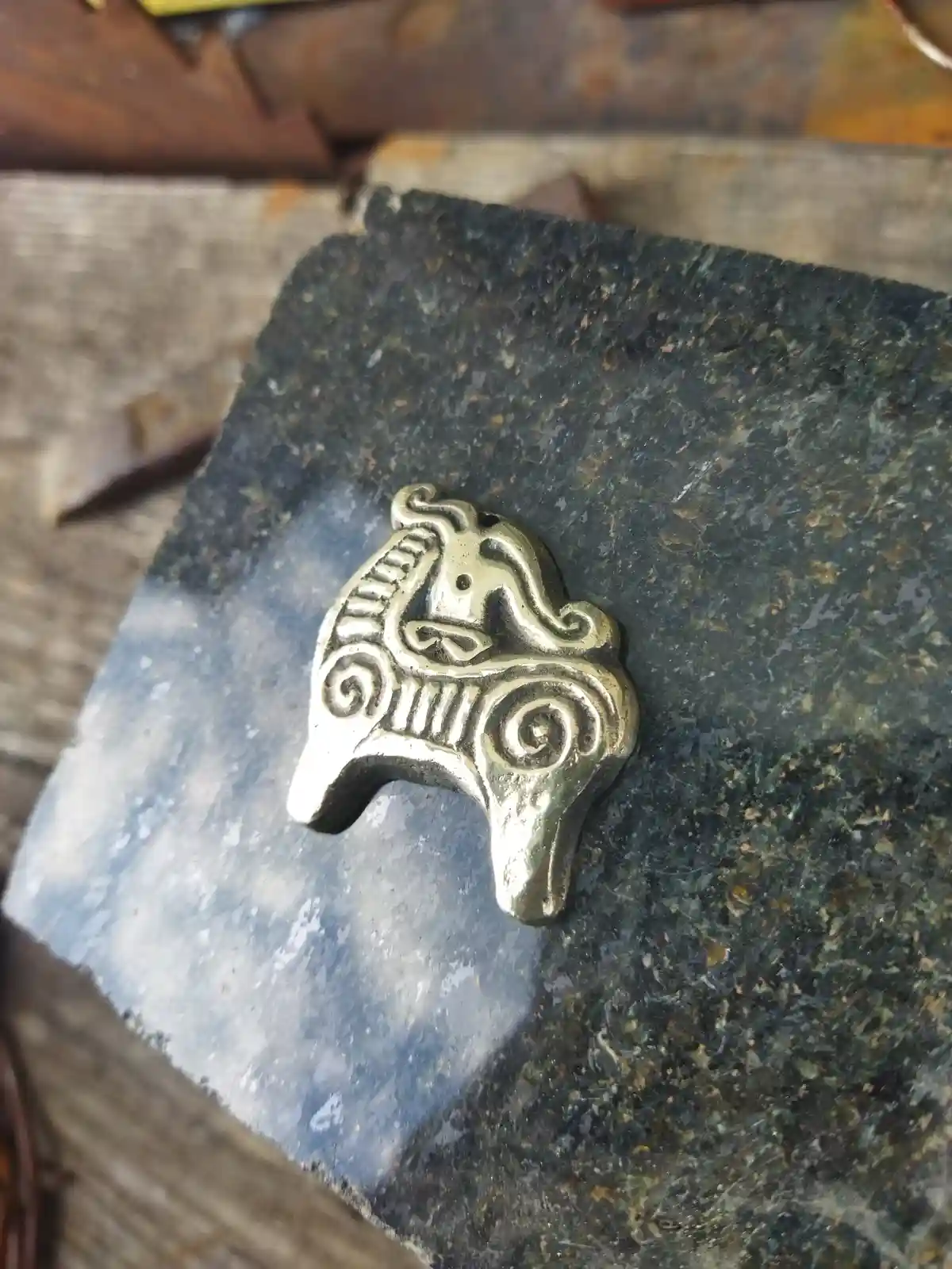 A small, ornate, metallic horse-shaped charm with swirling patterns rests on a dark, speckled stone surface, likely a…