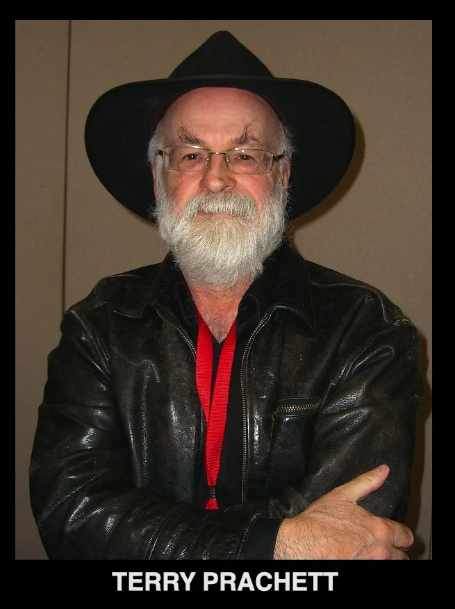 A man with a white beard, glasses, and a black wide-brimmed hat stands with arms crossed, wearing a black leather jacket…