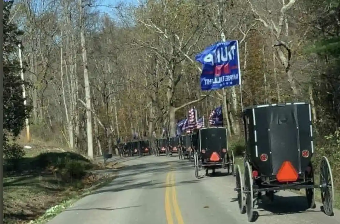 A line of black Amish-style horse-drawn buggies travels down a tree-lined road, with a large blue 'TRUMP 2024 PRESIDENT'…