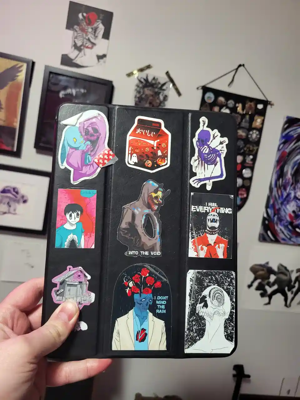 A hand holds a black tablet case covered in various dark, surreal, and emo-style stickers featuring skulls, monsters, and…