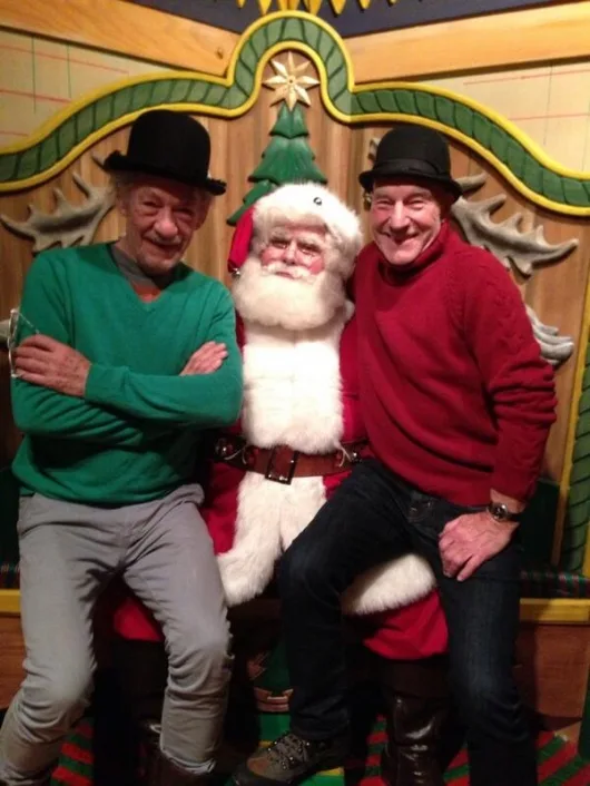 Two men in bowler hats flank a seated Santa Claus figure against a festive backdrop with a Christmas tree and antler…