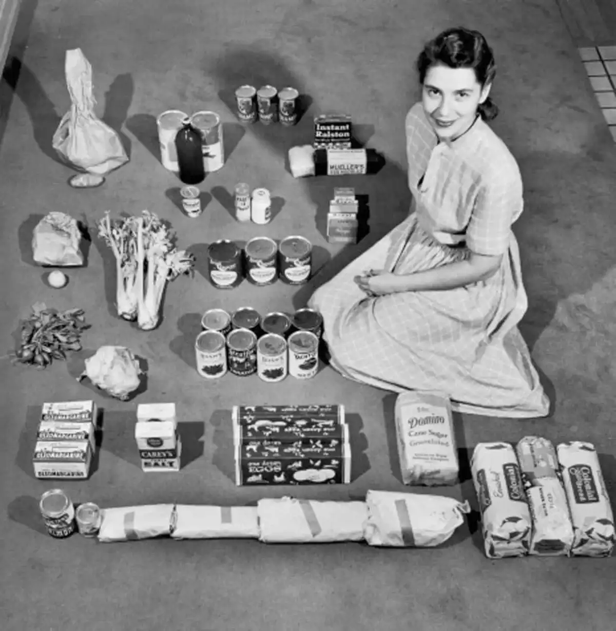 A vintage black-and-white photo shows a smiling woman in a checkered dress kneeling beside a large, meticulously arranged…