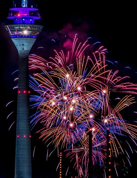 A tall, illuminated tower stands against a dark night sky, with vibrant fireworks exploding in colorful bursts around its…